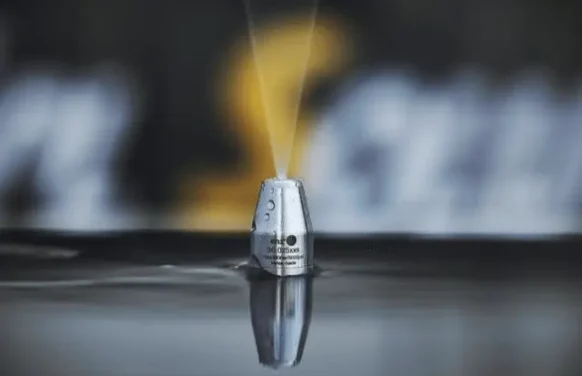 Close-up of a metal nozzle, spraying water in a vertical stream. The nozzle is partially submerged in a dark liquid surface, highlighting the expertise of plumbing contractors Pasco County FL. The background is blurred, featuring indistinct yellow and white shapes.