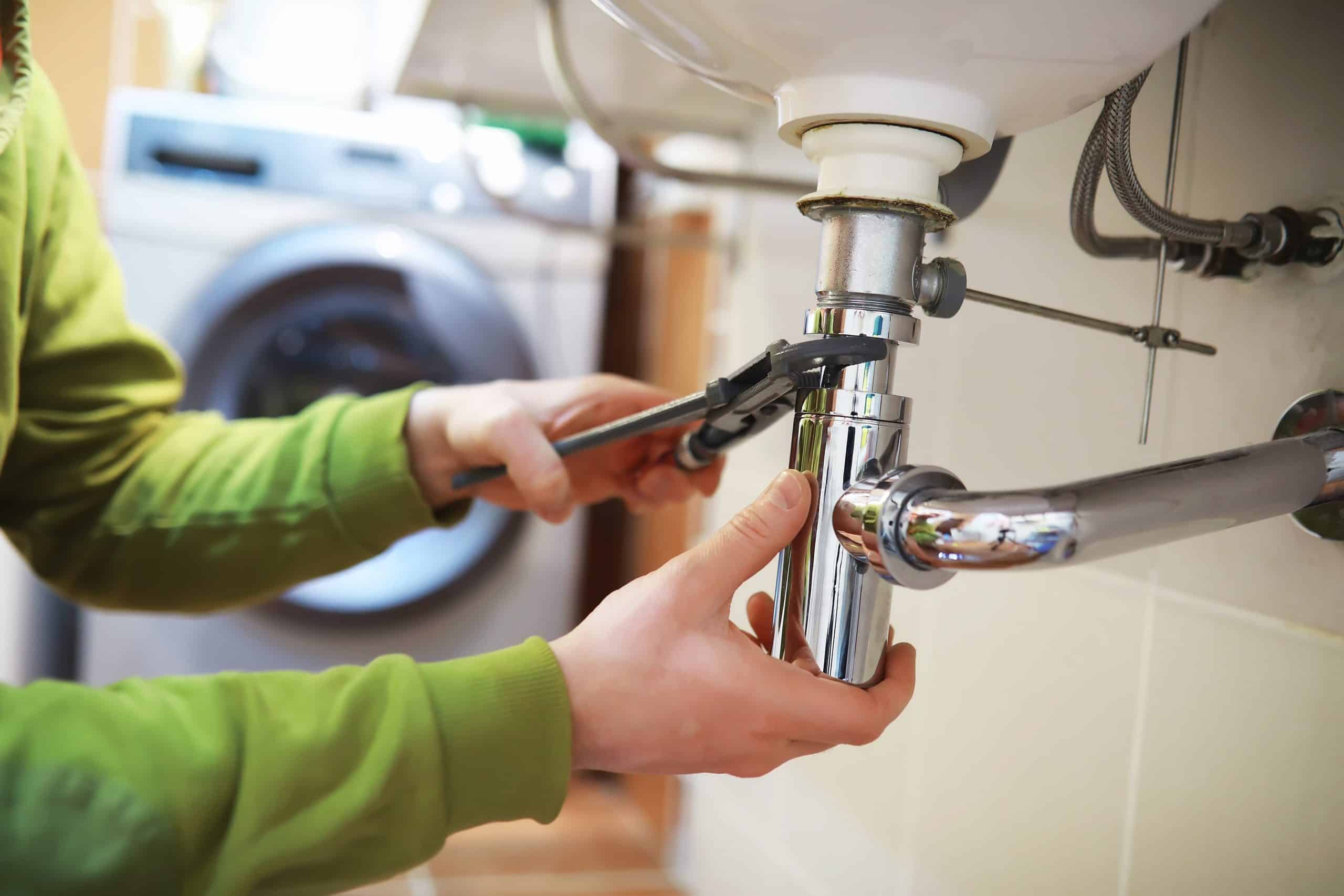 A person in a green shirt uses a wrench to tighten a pipe under a white sink. A washing machine is visible in the background, indicating a laundry or utility room setting. The focus is on the hands and plumbing work.