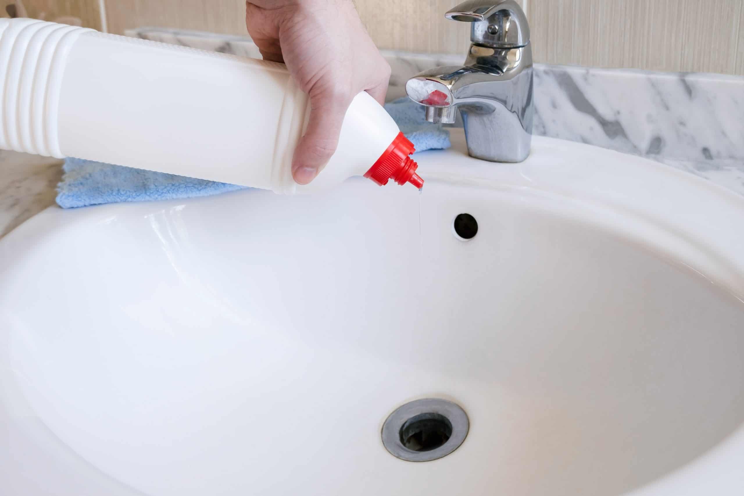 A person is pouring cleaning liquid from a white bottle with a red spout into a bathroom sink. A silver faucet and blue cloth are visible on the marble counter.