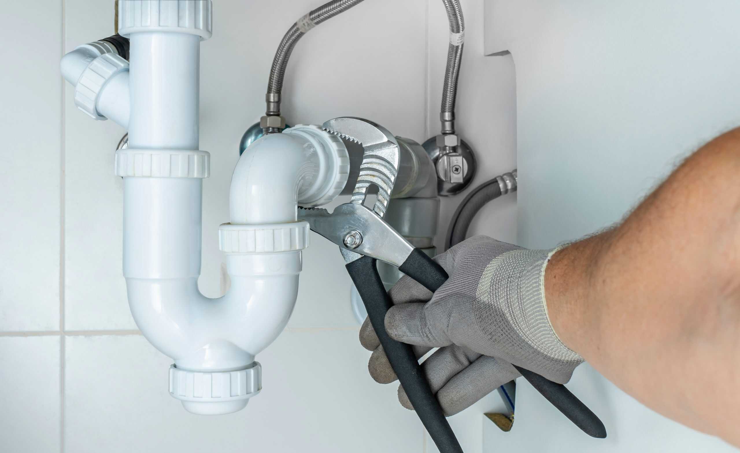 Close-up of a person’s gloved hand using pliers to tighten the pipes under a sink. The plumbing includes white PVC components and metal fittings, set against white tiled walls.