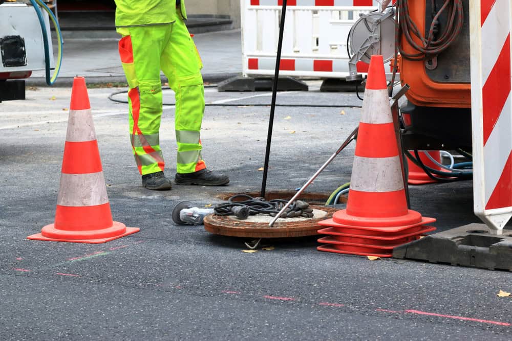 A construction worker in fluorescent gear stands near a manhole on a street. Orange traffic cones surround the area. Various equipment is visible, with a work vehicle partially seen on the right.