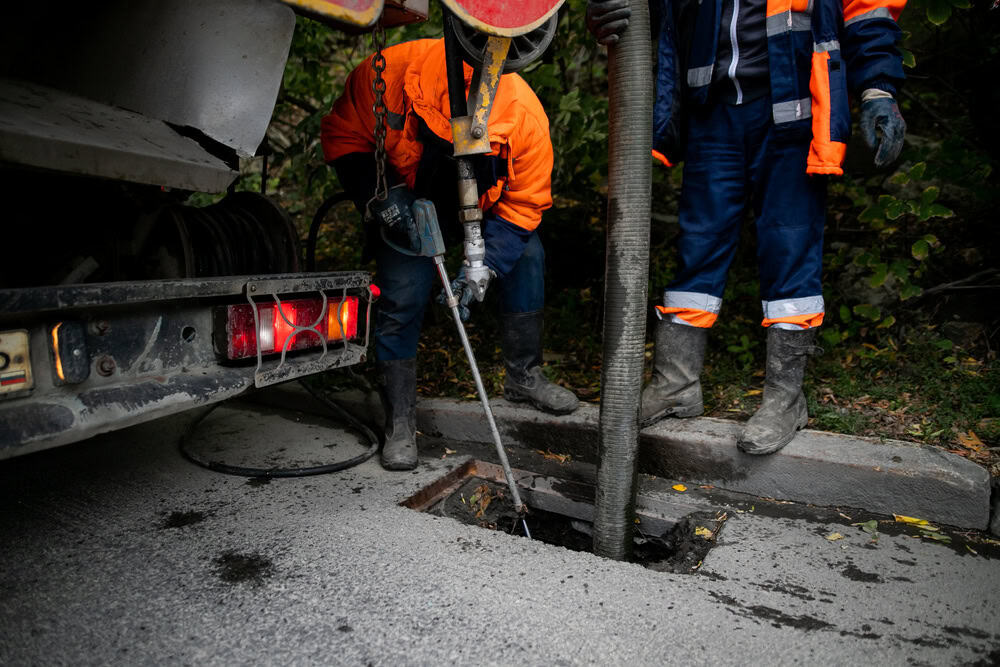 Two workers in orange and blue uniforms operate equipment from a truck to clean a sewer. One holds a hose inserted into a drain, while the other assists. They stand on a paved road with surrounding greenery.