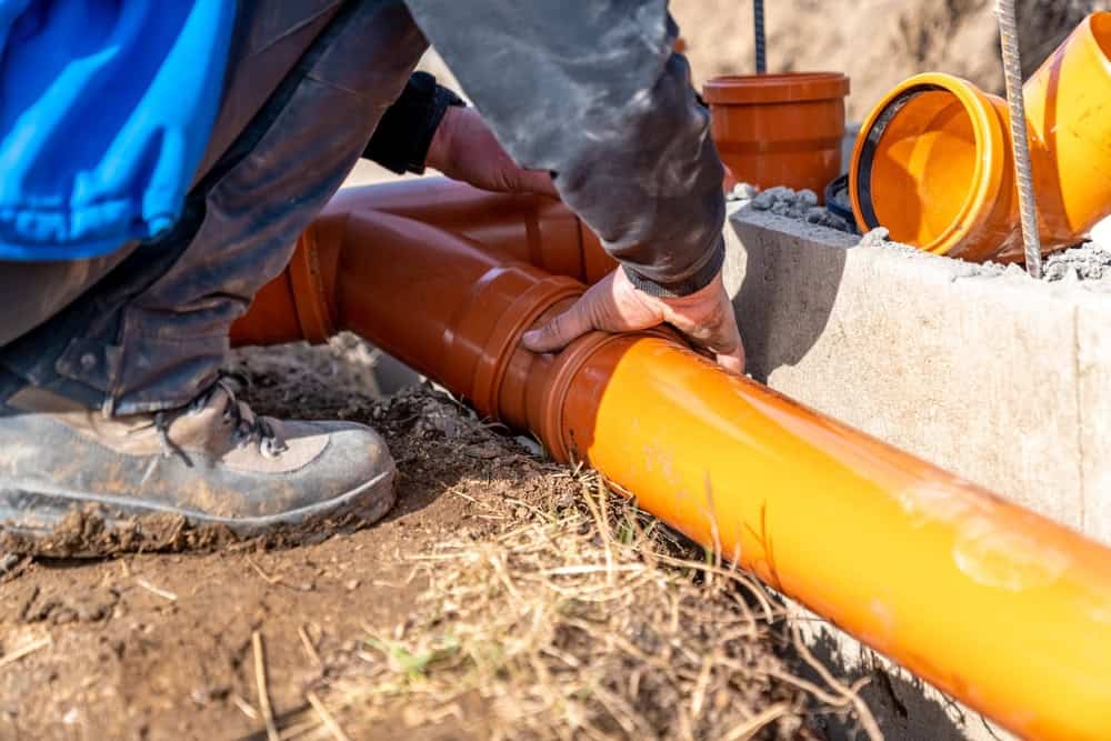 A person wearing a blue jacket and dark pants installs an orange drainage pipe into a fitting at a construction site. The ground is muddy, and various pipe components are scattered around.
