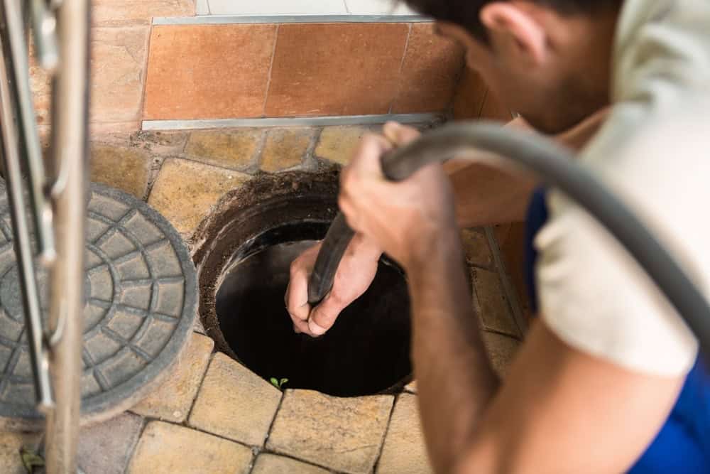 A person cleaning or inspecting a circular drainage hole with a tool on a tiled floor. A round manhole cover is partially visible nearby. The scene suggests maintenance or repair work in progress.
