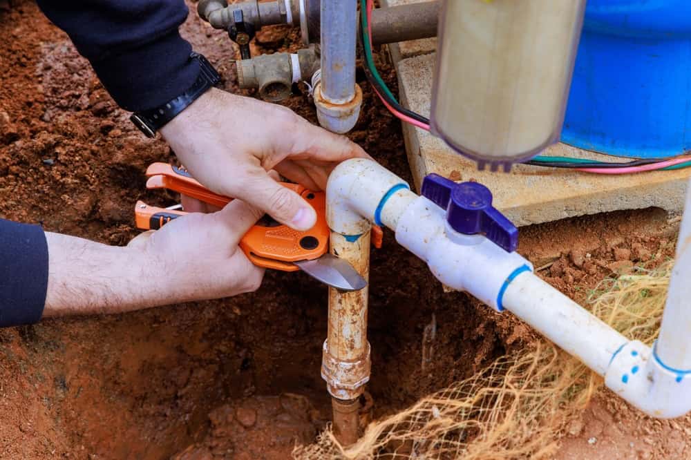A person uses an orange pipe cutter to work on white PVC pipes in a dirt area. The pipes are connected to various fittings, including a blue valve. The ground is reddish-brown, and there are some exposed roots nearby.