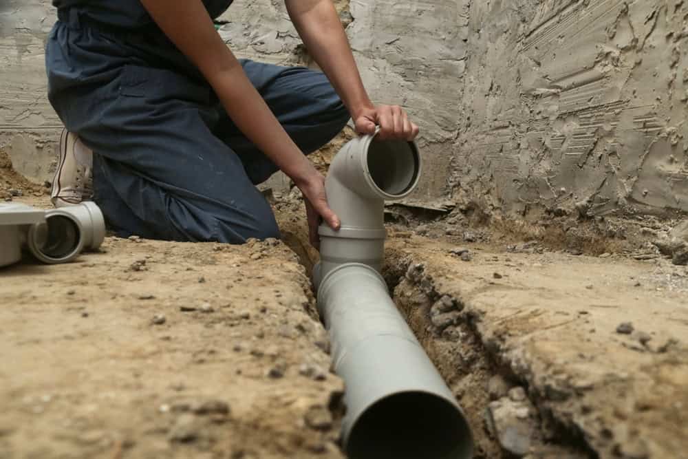 Person in overalls kneeling in a trench, assembling grey PVC plumbing pipes against a rough concrete wall. The individual is fitting an elbow joint to a straight pipe, surrounded by dirt and tools.