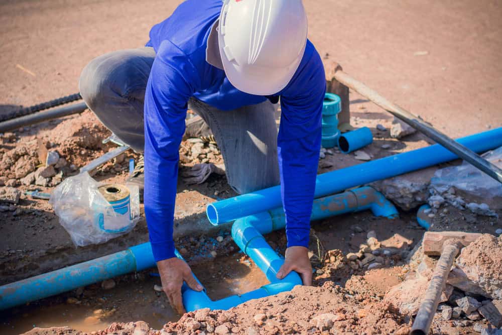 Worker wearing a white hard hat and blue shirt installs blue PVC pipes in a trench at a construction site. The area is surrounded by dirt and rocks, with speckled sunlight illuminating the scene.