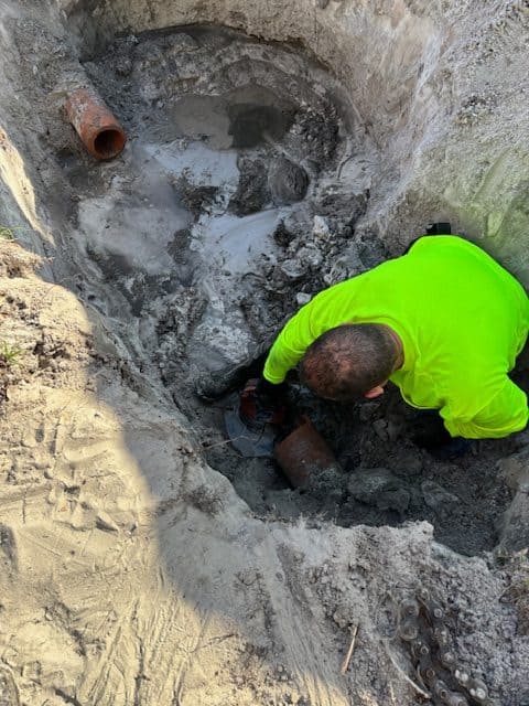 A worker in a neon yellow shirt is kneeling in a muddy trench, showcasing the expertise of Plumbing Contractors as he works with two exposed clay pipes surrounded by wet soil and water.