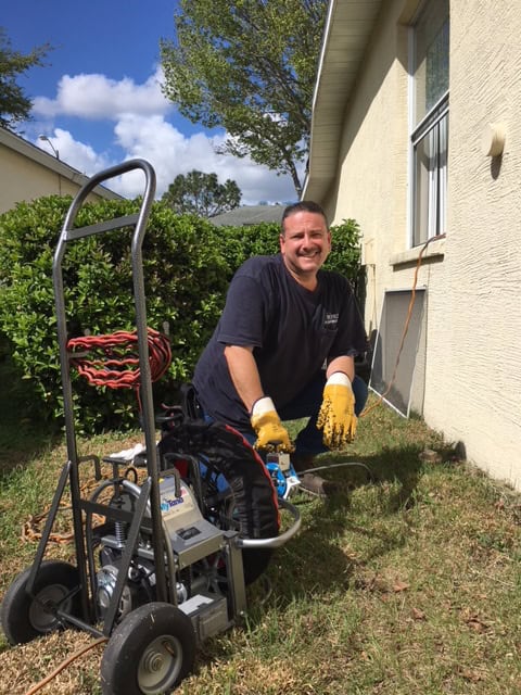 A smiling person wearing gloves kneels in a grassy yard beside a house, using outdoor plumbing company equipment with hoses and a reel. Green bushes, a window, and a blue sky with clouds complete the scene.