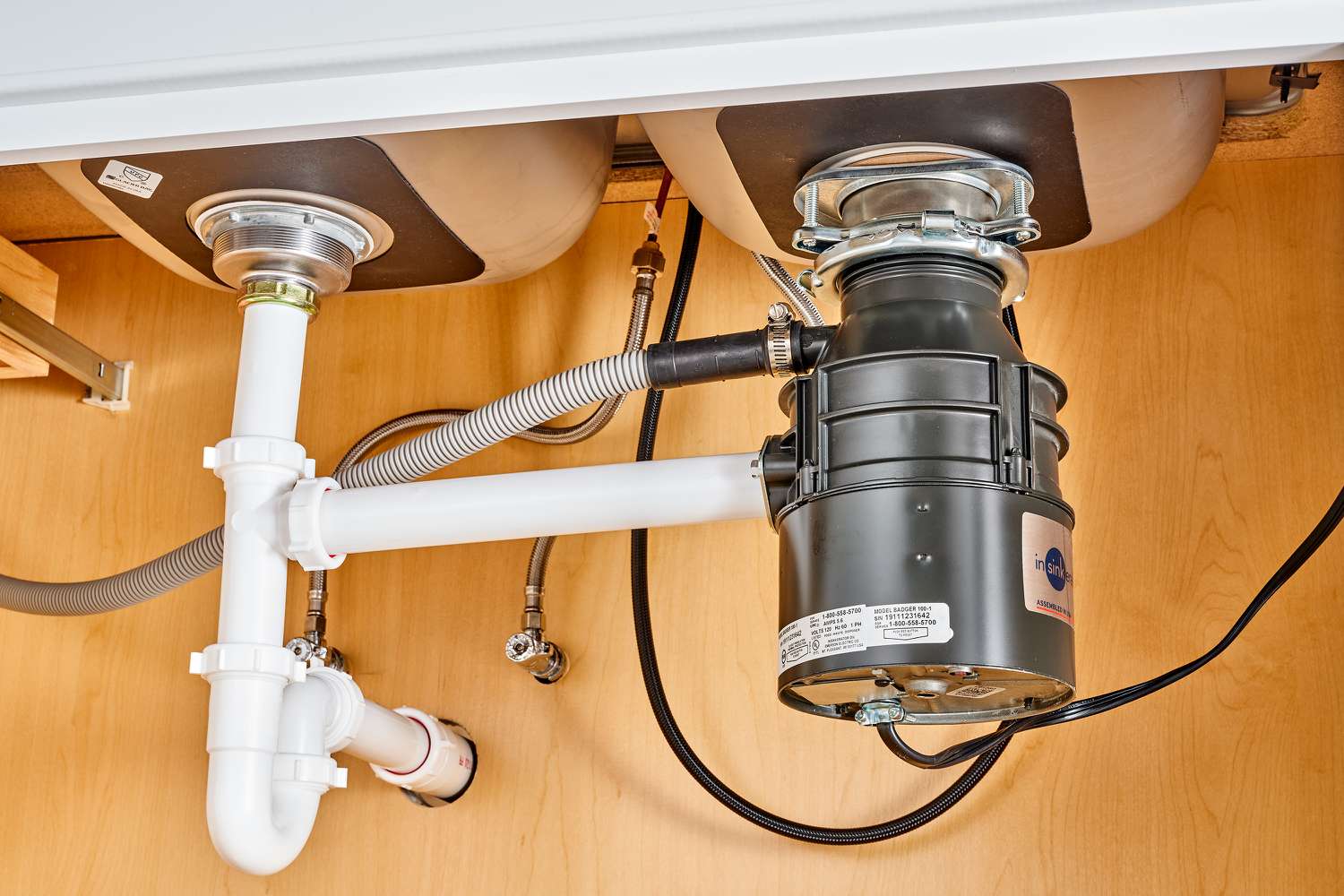 A garbage disposal installed under a double kitchen sink, connected to white PVC drain pipes and a dishwasher hose inside a clean wooden cabinet.