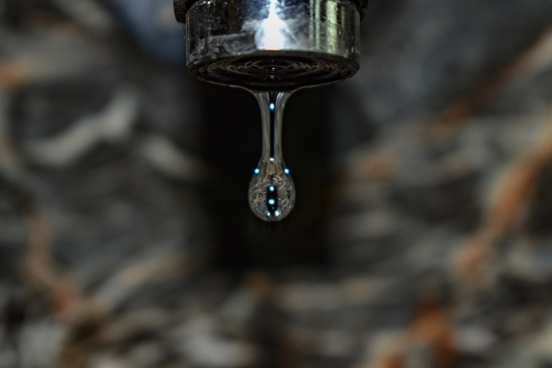 A close-up, moody shot of a metal faucet with a single droplet of water hanging from the tip. The droplet is sharply in focus, catching light and reflecting tiny highlights, while the background is softly blurred with dark, stone-like textures. The image emphasizes stillness, waste, and anticipation—capturing the exact moment before the drop falls.