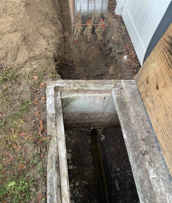 A concrete drainage channel lies open next to a house, surrounded by dirt and soil. Above, an excavator bucket hints at plumbing contractors from Pasco County FL working on construction or repairs. A wooden board partially covers one side, as work progresses seamlessly.
