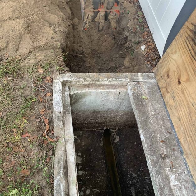A concrete drainage channel lies open next to a house, surrounded by dirt and soil. Above, an excavator bucket hints at plumbing contractors from Pasco County FL working on construction or repairs. A wooden board partially covers one side, as work progresses seamlessly.