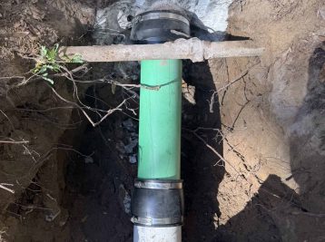 A section of green pipe connects to a white one in a dug trench, secured by metal clamps. Dirt and foliage surround the pipes, while a Plumbing Contractor from Pasco County, FL, ensures everything's in place; a brown boot peeks into the frame at the bottom right.