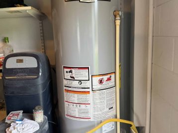 A gray water heater resides in the utility room, featuring a yellow gas line and various labels. Nearby shelves hold several items. If you're in need of expertise, Plumbing Contractors Pasco County FL offer reliable service to ensure everything flows smoothly.