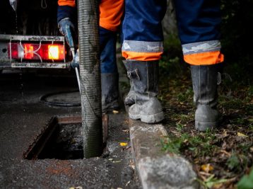 Worker in protective gear uses a large hose to clean a street drain. They are standing near a utility truck with bright lights. The ground is wet, and there are leaves scattered around.