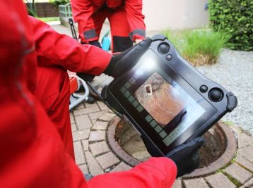 Person in red protective clothing holds a tablet displaying a sewer inspection image above an open manhole.